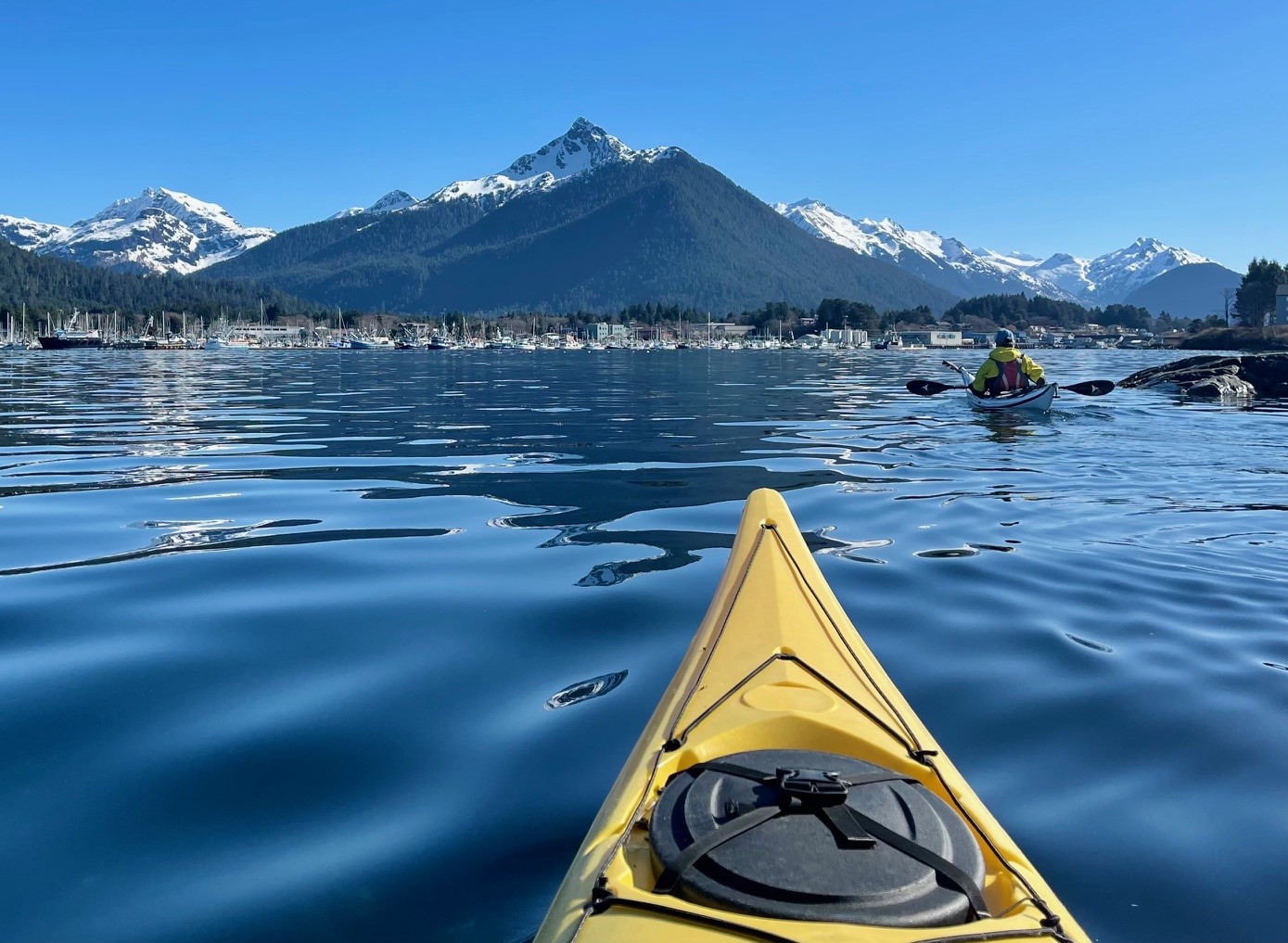 A kayaker crosses calm blue water toward Sitka and its snow-lined mountains.